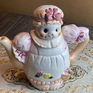 May include: A white ceramic teapot shaped like a girl wearing a pink dress and a pink and white hat. The teapot has a floral design and a pink bow on the dress.