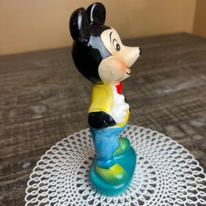 May include: A vintage ceramic figurine of Mickey Mouse wearing a yellow shirt, blue pants, and a red bow tie. The figurine is standing on a blue and green base.