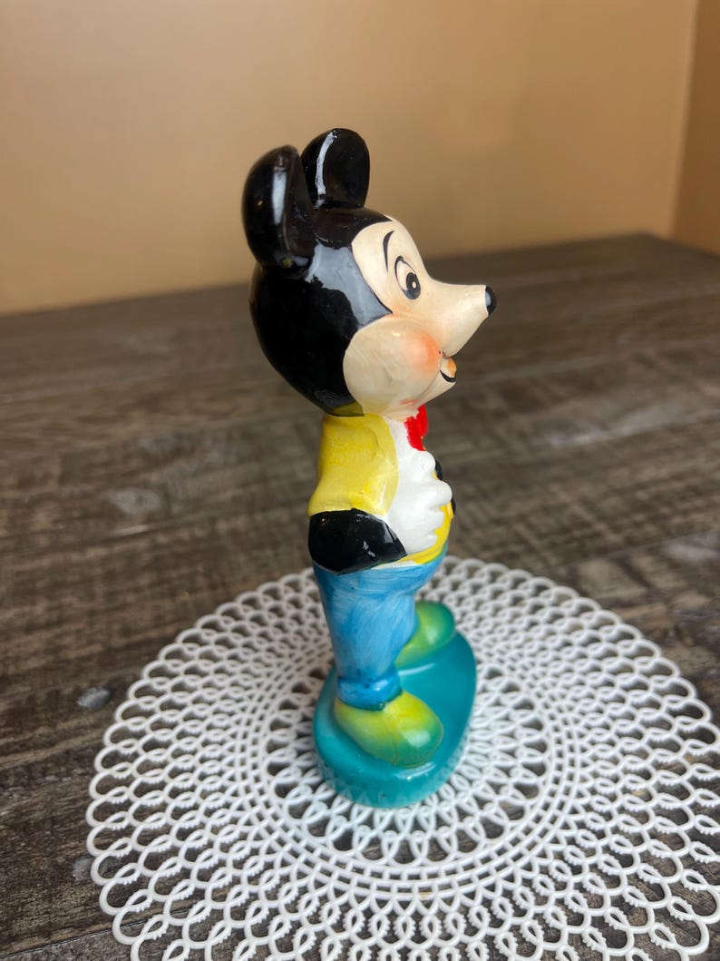May include: A vintage ceramic figurine of Mickey Mouse wearing a yellow shirt, blue pants, and a red bow tie. The figurine is standing on a blue and green base.