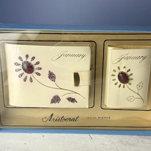 May include: A vintage Aristocrat Fifth Avenue desk calendar set. The set includes a cream-colored calendar and notepad, each adorned with a floral design and the month of "January". The set is housed in a blue box.