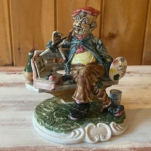 May include: A ceramic figurine of an artist seated on a bench, smoking a pipe, and holding a palette. The artist wears a red beret, a blue jacket, and brown pants. A bottle and paint can are nearby. The base is green.