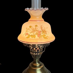 May include: A vintage-style table lamp with a floral design on the shade. The lamp features a brass base and a clear glass chimney. The shade is cream-colored with pink and yellow flowers.