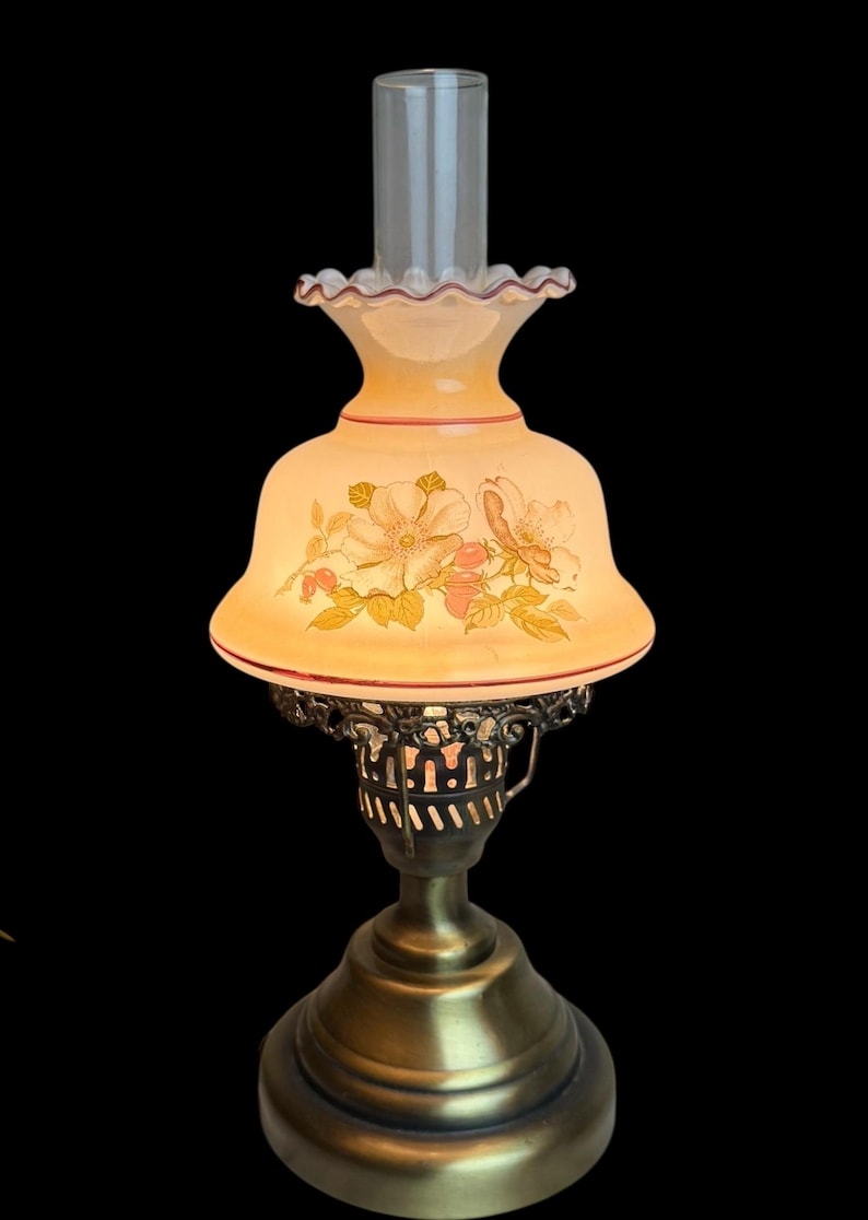 May include: A vintage-style table lamp with a floral design on the shade. The lamp features a brass base and a clear glass chimney. The shade is cream-colored with pink and yellow flowers.