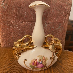 May include: A white ceramic vase with gold accents and a scene of a couple in a garden. The vase has two gold bird handles.