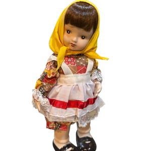 May include: A vintage porcelain doll with brown hair and a yellow headscarf. The doll wears a colorful floral print dress with a white apron and a red ribbon. Black shoes complete the outfit. A collectible doll.