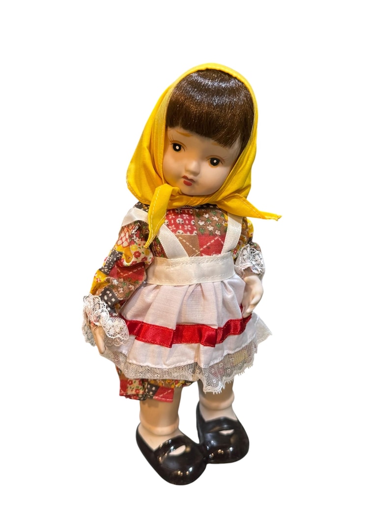 May include: A vintage porcelain doll with brown hair and a yellow headscarf. The doll wears a colorful floral print dress with a white apron and a red ribbon. Black shoes complete the outfit. A collectible doll.