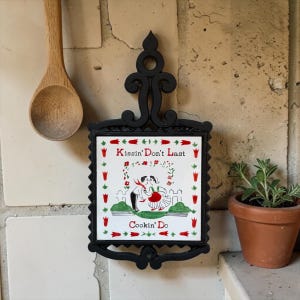 May include: Black cast iron trivet with a ceramic tile featuring a couple kissing, surrounded by red tulips. The text on the tile reads "Kissin' Don't Last Cookin' Do". A wooden spoon hangs to the left.