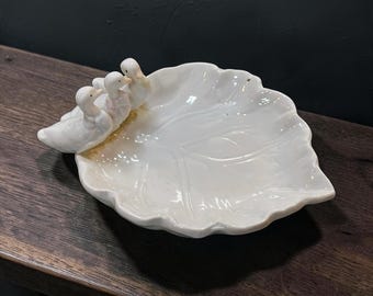 May include: A white ceramic dish shaped like a leaf with three small duck figurines attached to one side. The dish features a glossy finish and embossed vein details on the surface, while the ducks are sculpted with smooth, rounded forms.