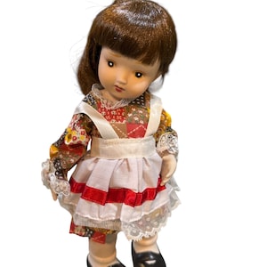 May include: A vintage porcelain doll with brown hair and a detailed face. The doll wears a colorful patchwork dress with a white apron and red accents. Black shoes complete the outfit. A collectible item.