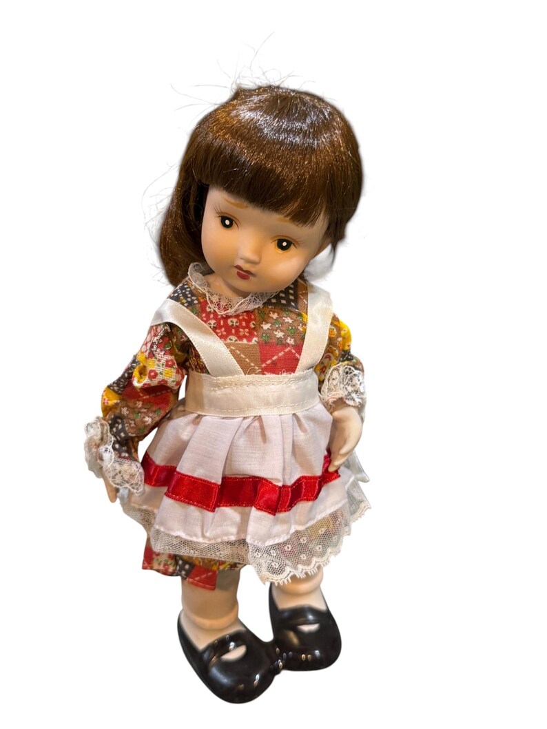 May include: A vintage porcelain doll with brown hair and a detailed face. The doll wears a colorful patchwork dress with a white apron and red accents. Black shoes complete the outfit. A collectible item.
