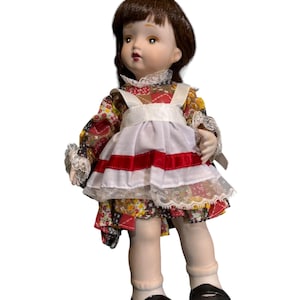 May include: A porcelain doll with brown hair and a floral patterned dress. The doll wears a white apron with red stripes and black shoes. The doll's dress has lace trim and the doll is posed with a slight tilt.