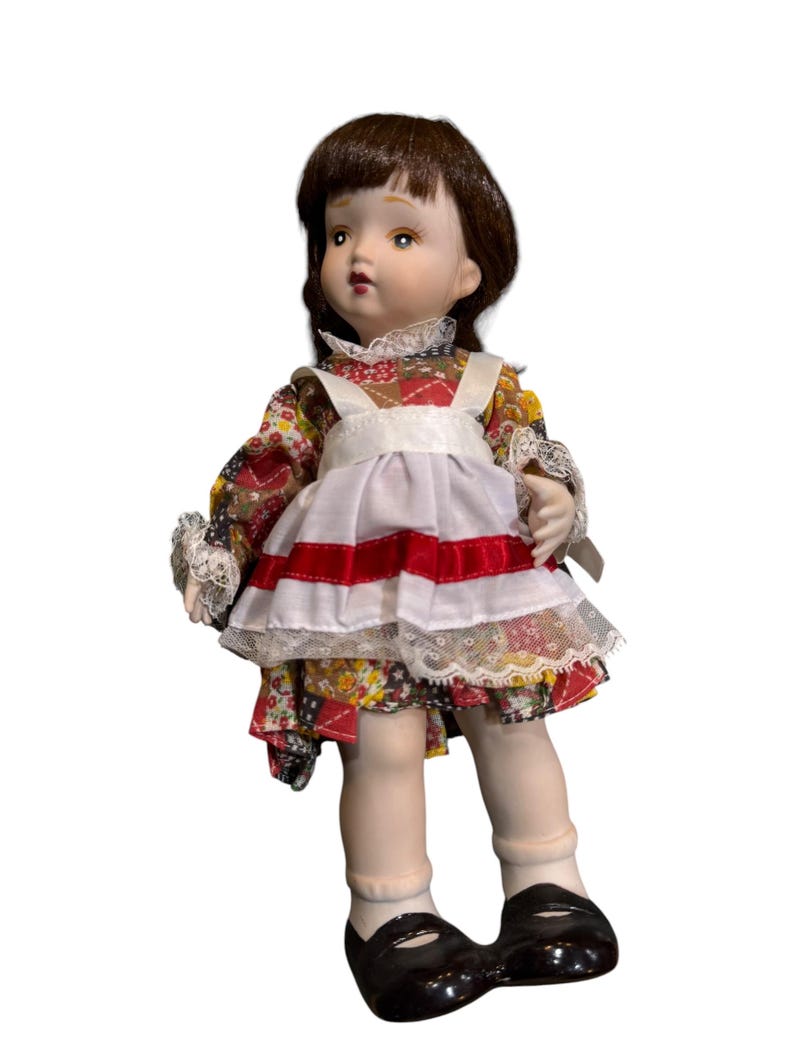 May include: A porcelain doll with brown hair and a floral patterned dress. The doll wears a white apron with red stripes and black shoes. The doll's dress has lace trim and the doll is posed with a slight tilt.