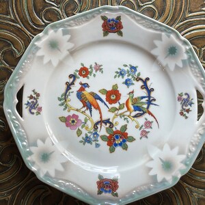 May include: A white porcelain plate with a scalloped edge and two handles. The plate features a floral design with two colorful birds and a green and white border.