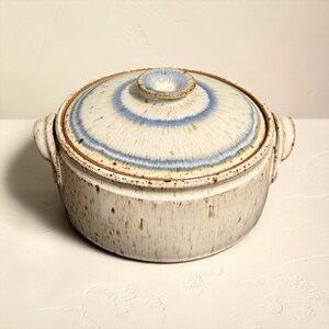 May include: A ceramic lidded casserole dish with a speckled beige exterior and a lid featuring concentric blue and white rings. The dish has two side handles and a small knob on the lid.