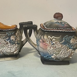 Op de afbeelding: Een bruine en witte keramische theepot en roomkan set met een drakenontwerp. De theepot heeft een deksel met een bruine knop. De roomkan heeft een handvat.