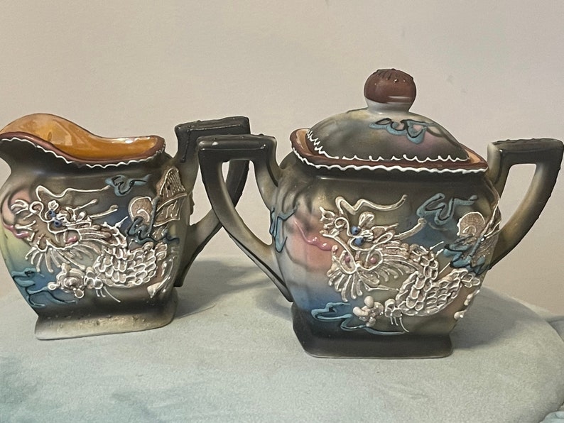 Op de afbeelding: Een bruine en witte keramische theepot en roomkan set met een drakenontwerp. De theepot heeft een deksel met een bruine knop. De roomkan heeft een handvat.