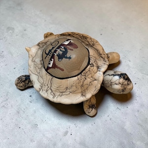 May include: A ceramic turtle figurine with a cream-colored body featuring fine black crackle patterns. The shell has a smooth, circular tan center medallion decorated with a dark brown and black graphic illustration of a musician playing a piano.