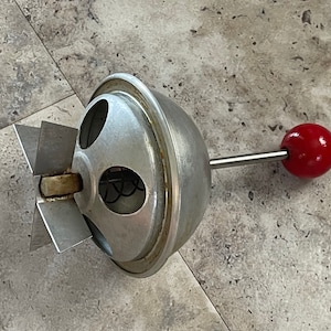 May include: A vintage, hand-cranked egg beater with a silver-colored metal body and a red wooden handle. The beater features a blade-like design and is likely used for mixing ingredients in cooking. The beater measures approximately 10 inches in length.