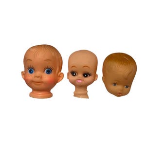 May include: Three plastic doll heads with painted features. The heads are all different, with one having blue eyes and rosy cheeks, one with brown eyes and pink lips, and one with brown eyes and a neutral expression.
