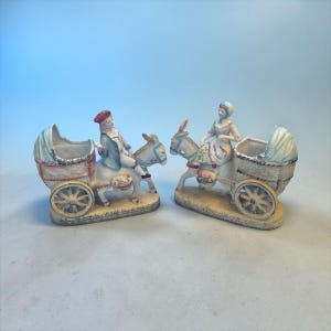 May include: Two ceramic figurines depicting people riding donkeys pulling baby carriages. The figures are dressed in period clothing, with the carriages featuring a light blue canopy. The figurines are painted in pastel colors and sit on a beige base.