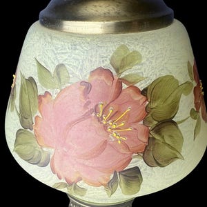 Puede incluir: Pantalla de l&aacute;mpara decorativa con dise&ntilde;o floral. La pantalla es de color verde p&aacute;lido con una superficie texturizada. Grandes flores rosas con hojas verdes est&aacute;n pintadas en la pantalla. La l&aacute;mpara tiene una base plateada y una parte superior de lat&oacute;n.