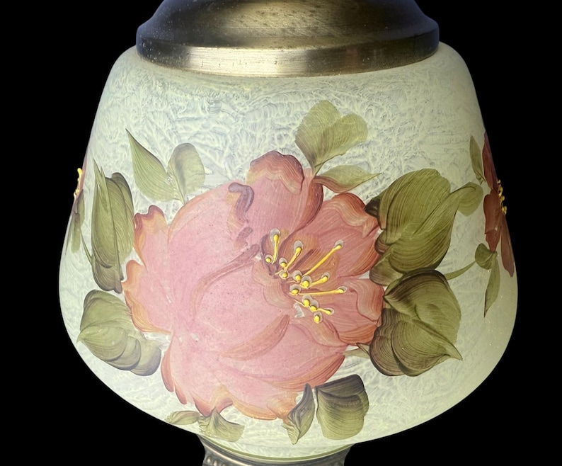 Puede incluir: Pantalla de l&aacute;mpara decorativa con dise&ntilde;o floral. La pantalla es de color verde p&aacute;lido con una superficie texturizada. Grandes flores rosas con hojas verdes est&aacute;n pintadas en la pantalla. La l&aacute;mpara tiene una base plateada y una parte superior de lat&oacute;n.
