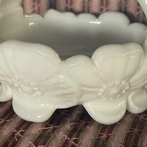 May include: White milk glass bowl with a scalloped rim and floral design. The bowl has a handle and sits on three feet.
