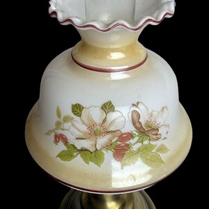 May include: A vintage-style lamp with a white glass shade featuring a floral design with white and brown flowers, green leaves, and red berries. The shade has a ruffled edge with a maroon trim, and sits atop a brass-colored base.