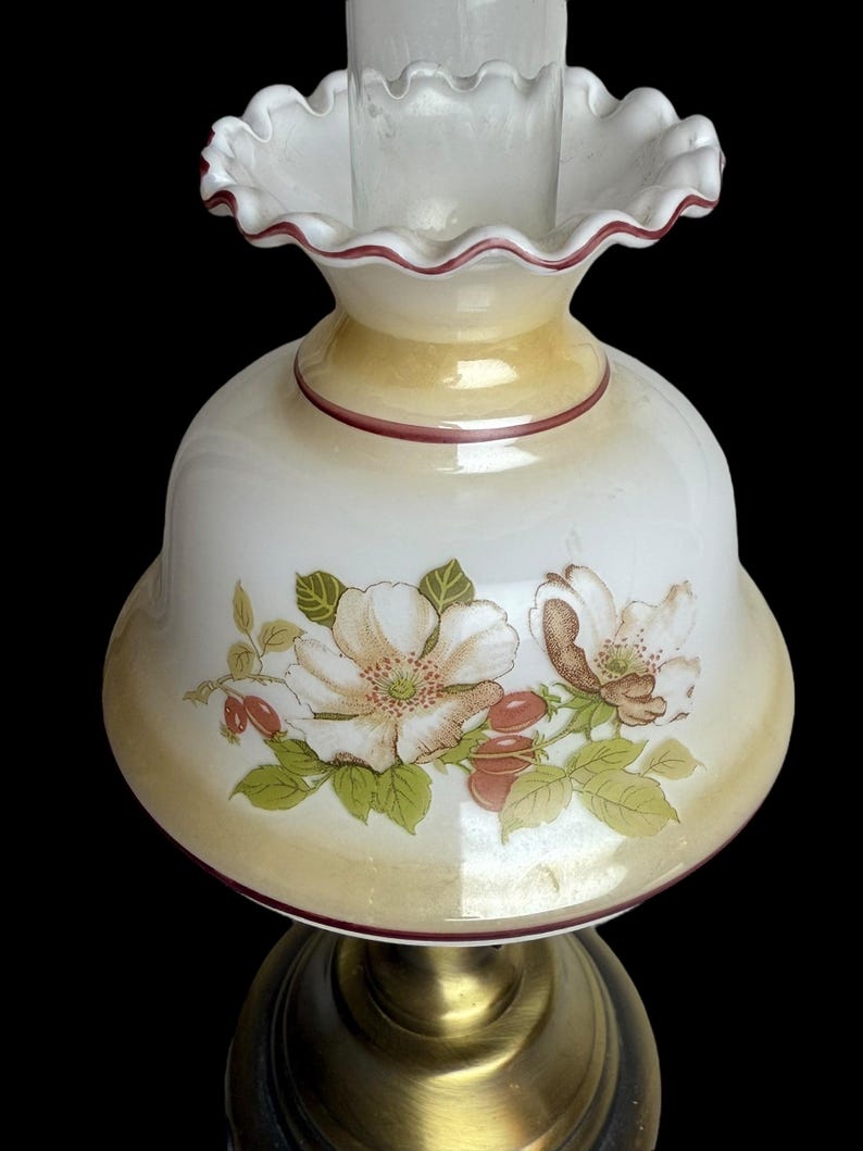 May include: A vintage-style lamp with a white glass shade featuring a floral design with white and brown flowers, green leaves, and red berries. The shade has a ruffled edge with a maroon trim, and sits atop a brass-colored base.
