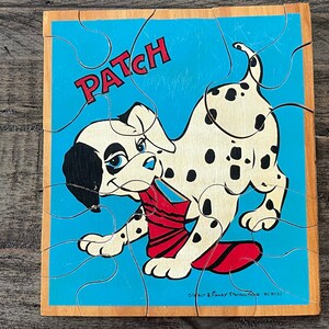 May include: Wooden jigsaw puzzle featuring a Dalmatian puppy named Patch. The puzzle has a blue background with the word "PATCH" in red. The puppy is white with black spots and is holding a red object in its mouth.