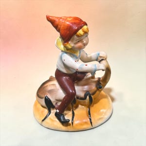 May include: A ceramic figurine depicting a small figure wearing a red pointed hat, a white shirt with red dots, and brown pants, riding on the back of a large tan beetle with black legs, all mounted on a circular base with a yellow-orange finish.
