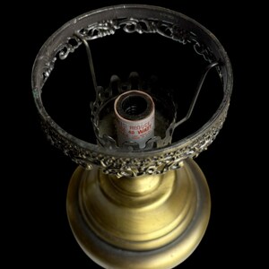 May include: A vintage brass lamp base with an ornate, filigree shade holder. The base has a round, pedestal design. The lamp is shown against a black background. The lamp is designed to hold a 40-watt bulb.