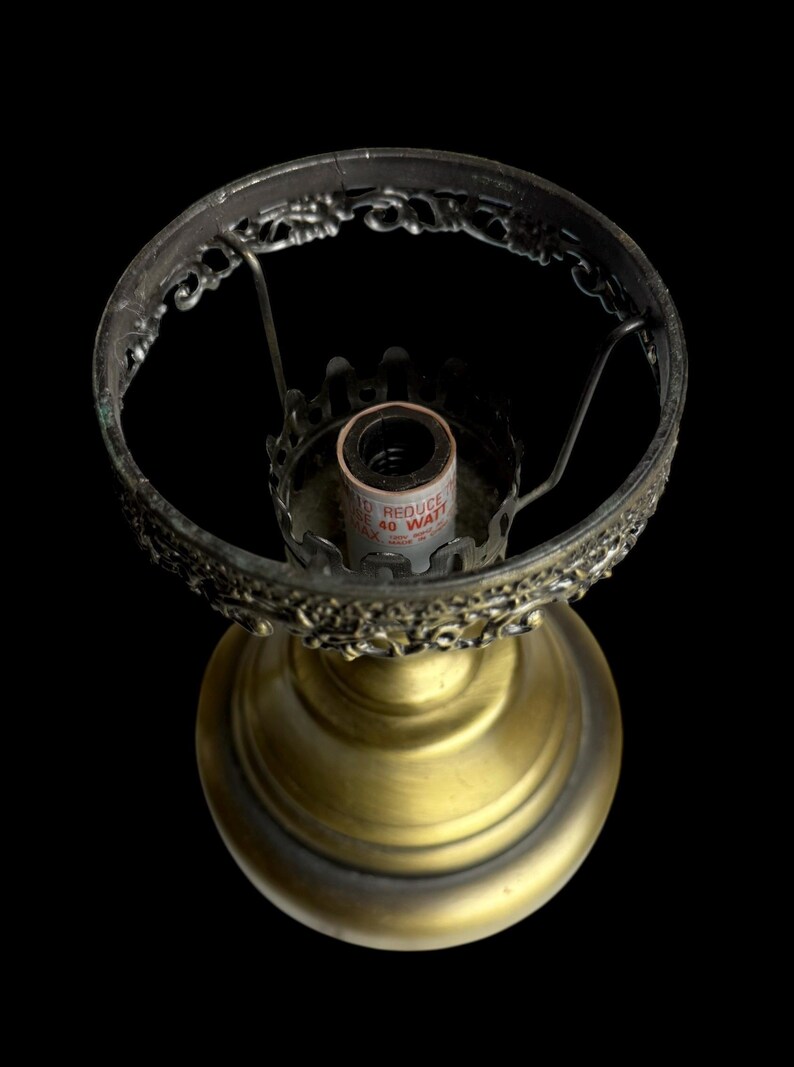 May include: A vintage brass lamp base with an ornate, filigree shade holder. The base has a round, pedestal design. The lamp is shown against a black background. The lamp is designed to hold a 40-watt bulb.