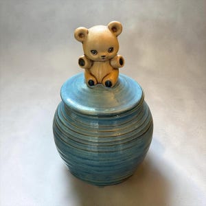 May include: A round, blue ceramic jar with a glossy finish and horizontal ribbed texture. The lid features a sculpted, light brown teddy bear figurine sitting on top, serving as a handle. The bear has dark eyes, a small nose, and dark paw accents.