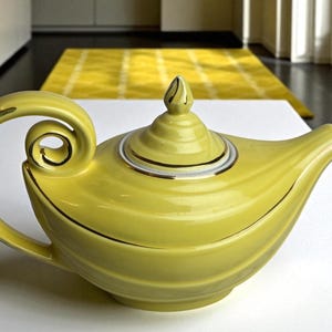 May include: A yellow ceramic teapot with a curved handle and spout. The teapot has a lid with a decorative finial and gold trim. The design features a swirl pattern on the body and handle. The teapot is a decorative kitchen item.