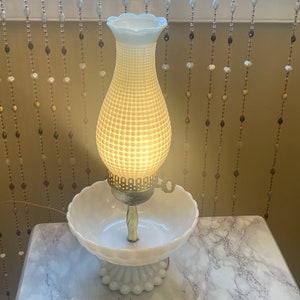May include: A white milk glass lamp with a textured shade and a white milk glass base. The lamp is lit and casting a warm glow.