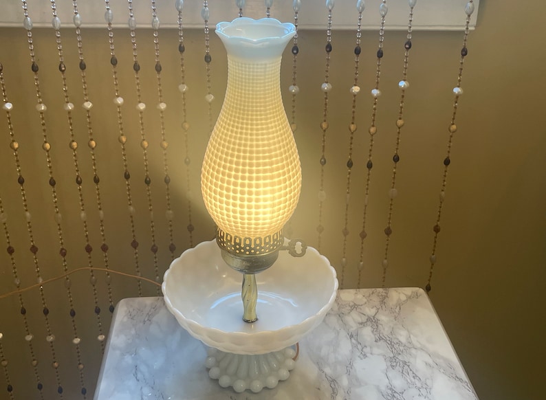 May include: A white milk glass lamp with a textured shade and a white milk glass base. The lamp is lit and casting a warm glow.