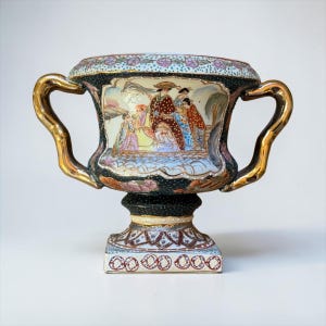 May include: A decorative ceramic urn featuring two gold-toned handles and a central panel depicting figures in traditional attire. The exterior is finished in a dark speckled pattern with floral accents, a flared rim, and a square pedestal base.