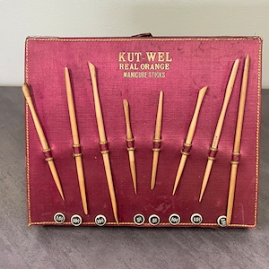 May include: A burgundy leather case with gold trim holds a set of eight wooden manicure sticks. The sticks are labeled with prices in cents. The case reads "KUT-WEL REAL ORANGE MANICURE STICKS".