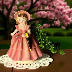 May include: A vintage ceramic figurine of a woman in a pink dress and hat, holding a black and yellow umbrella. The figurine is on a white lace doily, with a blurred background of pink flowers and greenery.