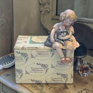 May include: A porcelain figurine of a young girl in a peach dress, holding a gray and white dog, sits atop a box. The box is cream-colored with floral patterns and the words "Treasured Memories" printed on it. A vintage aesthetic.