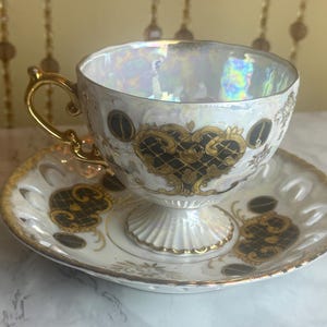 May include: A vintage teacup and saucer set. The cup and saucer are white with gold trim and accents. The cup has a gold handle and a pedestal base. The design includes black and gold filigree patterns.