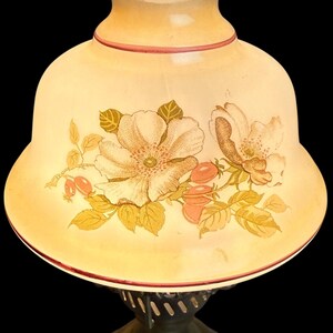 May include: A vintage-style table lamp with a cream-colored glass shade. The shade features a floral design in shades of peach, gold, and green. The base is a dark bronze color, and the lamp has a ruffled top edge.