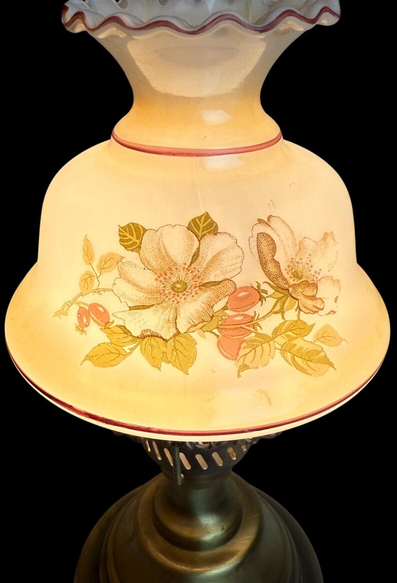 May include: A vintage-style table lamp with a cream-colored glass shade. The shade features a floral design in shades of peach, gold, and green. The base is a dark bronze color, and the lamp has a ruffled top edge.