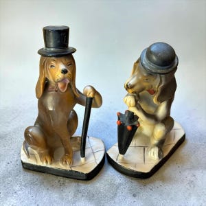 May include: Two ceramic dog figurines on rectangular bases. The left dog is brown, wearing a black top hat and holding a black cane. The right dog is light brown and white, wearing a grey bowler hat and holding a closed black umbrella with red accents.