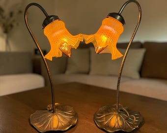 May include: Two matching table lamps featuring curved metal stems rising from textured, leaf-shaped bases. Each lamp is topped with a translucent, amber-colored glass shade shaped like a ruffled flower, emitting a warm, soft glow from within.