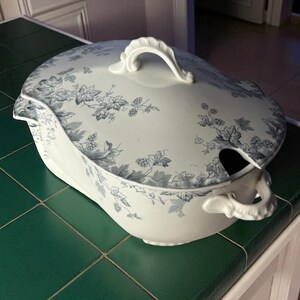 May include: Oval, white ceramic serving dish with a lid, featuring a blue floral pattern. The dish has a handle on the lid and on the side. A spoon slot is visible. A classic piece for serving food.