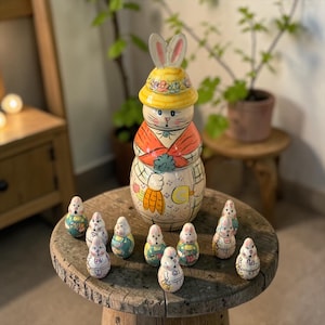 May include: A ceramic Easter bunny figurine with a yellow hat and carrot details, surrounded by smaller bunny figurines. The bunny figurines are painted with pastel colors and are arranged on a wooden table.