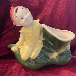 Puede incluir: Un macetero de cerámica con forma de hoja verde con una pequeña figura de elfo sentada encima. El elfo lleva un atuendo amarillo y verde y un gorro rojo.