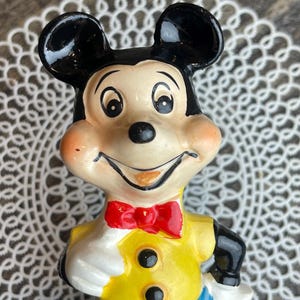 May include: A vintage ceramic figurine of Mickey Mouse. He is wearing a yellow shirt with two black buttons, a red bow tie, and blue pants. His face is painted with a friendly smile and large black eyes.